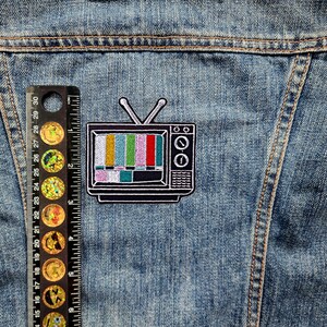 May include: A black and white embroidered patch of a vintage television with a colorful static screen. The patch is sewn onto a blue denim jacket.