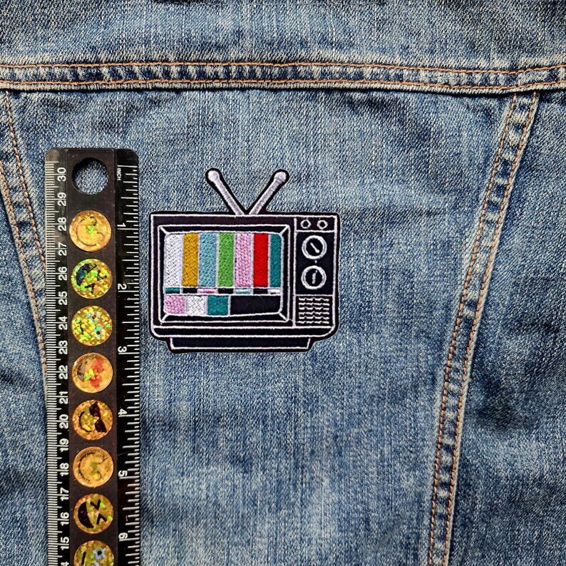 May include: A black and white embroidered patch of a vintage television with a colorful static screen. The patch is sewn onto a blue denim jacket.