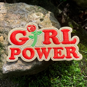 May include: A beige embroidered patch with the text "GIRL POWER" in red lettering. A red rose with a green stem is above the word "GIRL".
