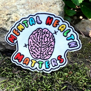 May include: Embroidered patch with the words "MENTAL HEALTH MATTERS" in rainbow colors. The patch features a pink brain graphic with black outlines. The patch is white with a green border.