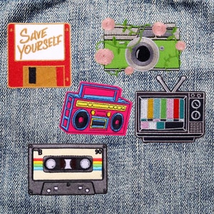 May include: Five embroidered patches on a denim background. The patches are a red floppy disk with the text "Save Yourself", a pink boombox, a green camera with pink flowers, a black and white television with a static screen, and a black cassette tape with rainbow stripes.