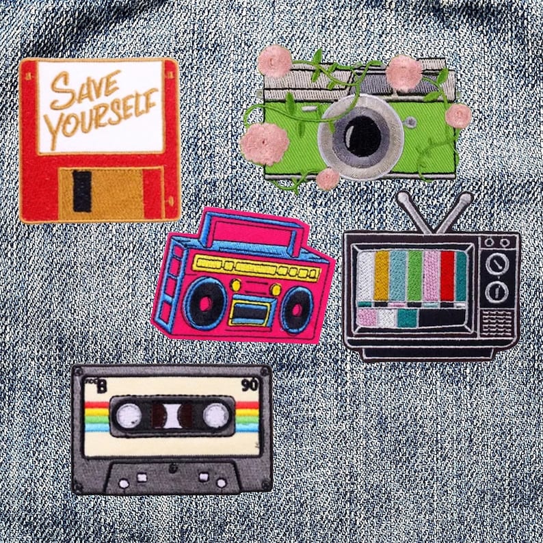 May include: Five embroidered patches on a denim background. The patches are a red floppy disk with the text "Save Yourself", a pink boombox, a green camera with pink flowers, a black and white television with a static screen, and a black cassette tape with rainbow stripes.
