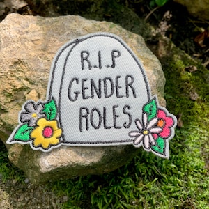 Peut inclure: Un patch brodé gris en forme de pierre tombale avec le texte "RIP Gender Roles" en noir. Le patch est décoré de fleurs brodées colorées.