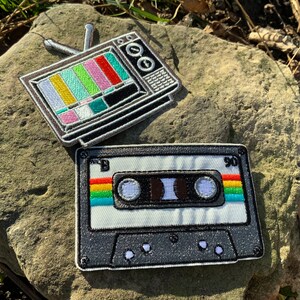 May include: Two embroidered patches, one of a vintage television with a test pattern on the screen and the other of a cassette tape with rainbow colored stripes on the sides.