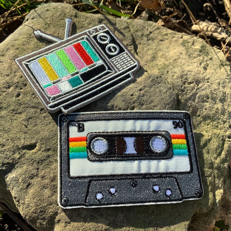 May include: Two embroidered patches, one of a vintage television with a test pattern on the screen and the other of a cassette tape with rainbow colored stripes on the sides.