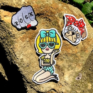Puede incluir: Tres parches bordados para planchar. Un parche presenta un puño con la palabra "ROCK" en los nudillos. Otro parche presenta una chica de dibujos animados con cabello rubio, gafas de sol y un traje de baño azul. El tercer parche presenta un pug de dibujos animados con una bandana roja.