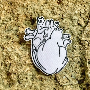 May include: A white patch featuring a heart shape with several cartoon cats inside. The cats are outlined in black, with a white background. The patch is on a textured, tan surface.