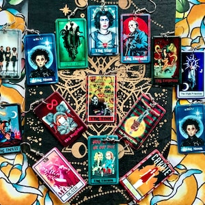 Puede incluir: Un conjunto de 12 pares de pendientes con personajes de películas de terror e imágenes de cartas del tarot. Los pendientes están hechos de madera y tienen una forma rectangular. Las imágenes incluyen personajes de las películas Carrie, El Resplandor, El Diablo, El Loco, El Mago, La Emperatriz, La Suma Sacerdotisa, La Estrella, El Ermitaño, Los Enamorados y El Diablo.