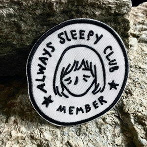 May include: White round patch with black embroidery. The patch says "Always Sleepy Club Member" with a cartoon face in the center.