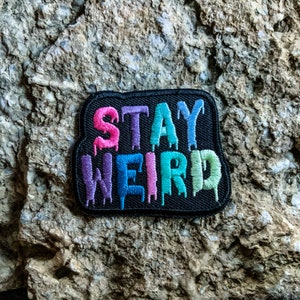 May include: A black patch with the words "STAY WEIRD" in rainbow dripping letters. The patch is made of fabric and has a black border.