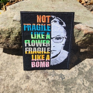 May include: A black embroidered patch with a portrait of Ruth Bader Ginsburg and the text "NOT FRAGILE LIKE A FLOWER FRAGILE LIKE A BOMB".