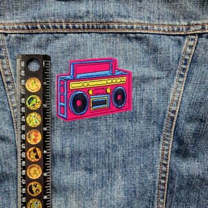 May include: A pink and blue retro boombox patch with yellow accents. The patch is iron-on and features a cassette tape player with a graphic design.