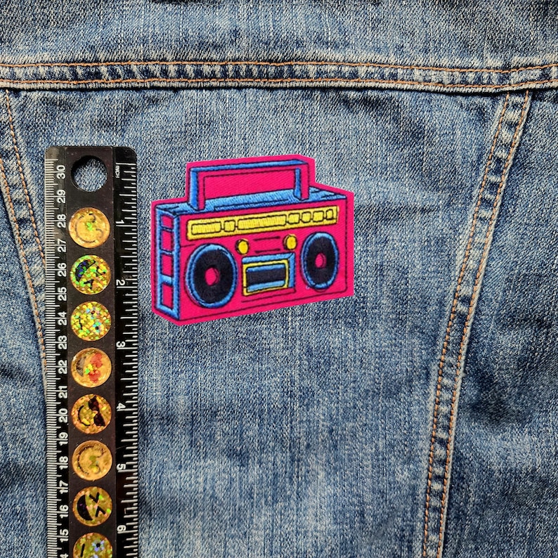 May include: A pink and blue retro boombox patch with yellow accents. The patch is iron-on and features a cassette tape player with a graphic design.