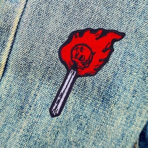 Peut inclure: Patch brodé rouge d'un crâne sur un bâton avec des flammes, sur un fond de denim bleu.