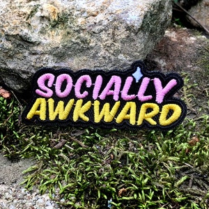 Puede incluir: Un parche bordado negro con el texto "SOCIALLY AWKWARD" en letras rosa y amarillo.