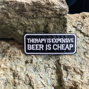 Op de afbeelding: Zwart-wit geborduurde patch met de tekst "Therapie is duur, bier is goedkoop".