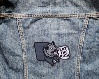 May include: A black and white embroidered patch of a cartoon opossum with its mouth open, saying "Eat the Rich" in a speech bubble. The patch is sewn onto a blue denim jacket.