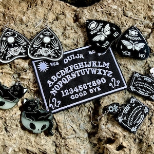 May include: A set of black and white earrings featuring a Ouija board design, a cat in a skull, and a moth with the words "Goodbye" and "Yes" on it. There are also two black and white Ouija board patches.