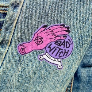 May include: Purple and pink embroidered iron-on patch with a hand holding a crystal ball that says "Bad Witch" on it.