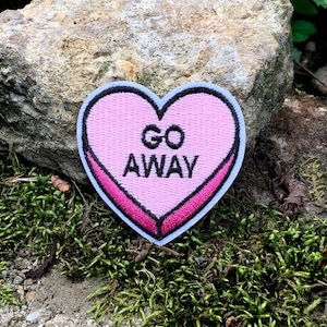 May include: A pink heart-shaped patch with the words "GO AWAY" in black lettering. The patch has a white border and a dark pink outline. The patch is set against a backdrop of moss and a rock.