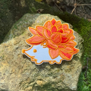 May include: A white embroidered hedgehog with a large orange and red flower on its back. The flower is made of multiple layers of petals and has a yellow outline.
