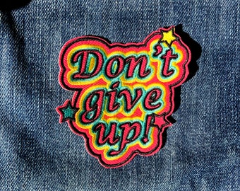 May include: A colourful embroidered patch with the text "Don't give up!" in a retro style. The patch has a red, yellow and green colour scheme with a black outline.