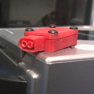 Może przedstawiać: Czerwony plastikowy adapter z dwoma wtyczkami umieszczony na srebrnej konsoli do gier. Konsola ma czarny uchwyt.