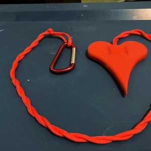 Può includere: Un oggetto rosso a forma di cuore con una piccola rientranza quadrata, collegato a un cordino intrecciato rosso. Un moschettone rosso è attaccato al cordino. Gli oggetti sono su una superficie blu scuro.