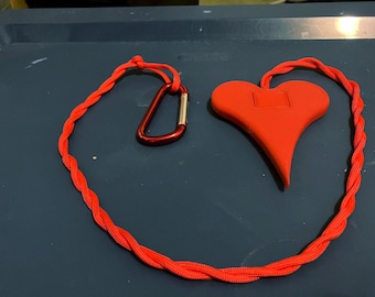 May include: A red heart-shaped object with a small square indentation, connected to a red braided cord. A red carabiner clip is attached to the cord. The items are on a dark blue surface.