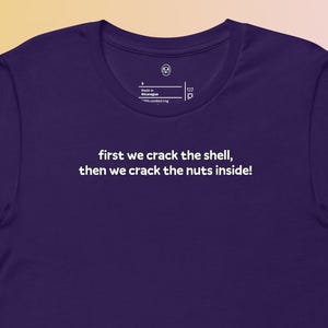 Könnte beinhalten: Ein dunkelviolettes T-Shirt mit dem weißen Aufdruck "first we crack the shell, then we crack the nuts inside!". Das Shirt hat kurze Ärmel und einen Rundhalsausschnitt. Der Hintergrund hat einen Farbverlauf von Gelb, Orange und Blau.