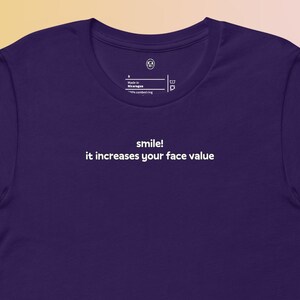 May include: A dark purple t-shirt with the text "smile! it increases your face value" printed in white. The shirt has a crew neck and short sleeves. The shirt is made of a soft material.