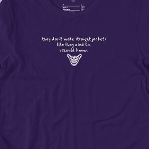 May include: A purple t-shirt with white text that reads "they don't make straight jackets like they used to. i should know." A smiling mouth graphic is below the text. The shirt is displayed on a flat surface with jeans and accessories.
