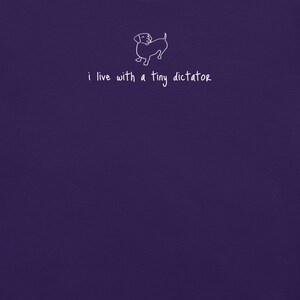 以下が含まれることがあります： 濃い紫色のTシャツ。白い線画で描かれたダックスフントの犬と「i live with a tiny dictator」の文字がプリントされています。犬のイラストはシンプルで、文字は手書き風です。