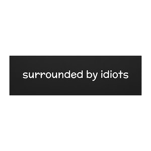 May include: A black rectangular sign with the phrase "surrounded by idiots" in white, sans-serif font. The sign is centered and the text is clearly legible against the dark background.