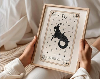 May include: A framed print of a black and white illustration of the Capricorn zodiac sign. The illustration features a goat with a fish tail and stars on a beige background. The frame is made of light wood and has a white mat.