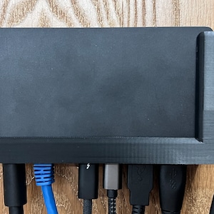 May include: A black plastic wall mount with a rectangular opening for a device and multiple ports on the bottom. The ports include a blue ethernet cable, a black USB-C cable, a silver USB-A cable, and a black HDMI cable.