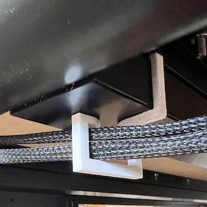 May include: White plastic cable management clip attached to the underside of a black desk, holding several black braided cables.