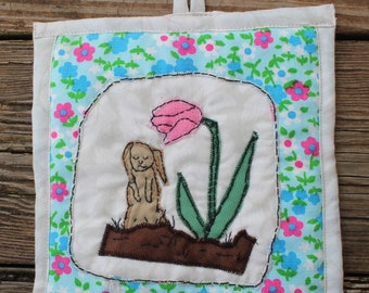 May include: A white fabric pot holder with a blue and pink floral border. The center features an embroidered image of a brown rabbit sitting next to a pink tulip with green leaves.