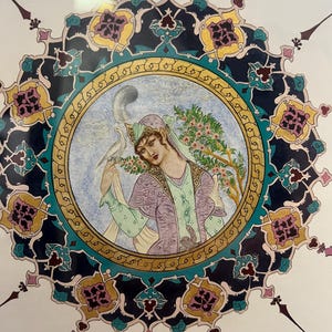May include: A circular painting featuring a figure in a lavender and green outfit with a feathered headpiece, holding a white bird. The central image is surrounded by an intricate, multi-colored border of dark blue, gold, and teal floral patterns.