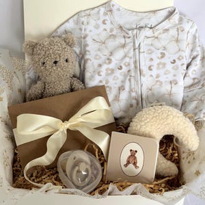 May include: A baby gift set in a white box, including a teddy bear, a floral-patterned onesie, a pacifier, a plush moon, and a small book with a teddy bear illustration. The gift box is tied with a cream-colored ribbon.