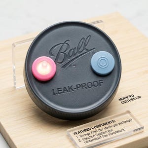 Puede incluir: Una tapa circular gris oscuro de la marca Ball a prueba de fugas con un filtro de jeringa rosa y un septo de inyección azul. La tapa está etiquetada como Modified Culture Lid e incluye una lista de componentes para intercambio gaseoso.
