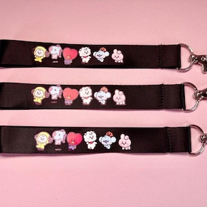 Puede incluir: Tres cordones de tela negra con un estampado gráfico repetitivo de seis personajes de dibujos animados coloridos. Cada cordón está rematado con un mosquetón de metal plateado unido a una anilla circular en un extremo de la cinta.