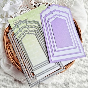 May include: A set of five silver metal die-cut frames with a decorative border. The frames are nested inside each other and have a scalloped edge. The frames are on a wicker surface.