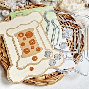 May include: A craft project featuring a beige spool-shaped die-cut with brown buttons, a metal die-cut, and a collection of small buttons. The items are arranged on a woven basket, suggesting a sewing or crafting theme.