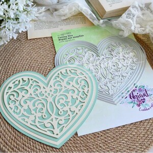 May include: A layered heart-shaped craft with intricate, openwork designs in white and a light blue backing. The craft is on a woven mat, with additional heart-shaped cutting dies and paper elements.