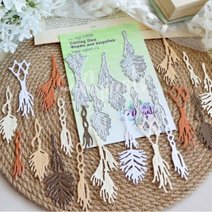 May include: A set of 10 metal die cuts in the shape of tassels with a feather design. The die cuts are in various shades of brown, beige, and white. The die cuts are arranged on a brown woven surface. The die cuts are for crafting and scrapbooking. The text on the packaging reads "Cutting Dies Forma Dlya vyrubki www.aglant.ru agl1998".