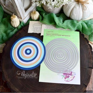 Puede incluir: Un proyecto de manualidades en papel en capas con círculos concéntricos en tonos azules, blancos y beige. Una plantilla troquelada con patrones circulares similares está junto a la pieza terminada. El proyecto está sobre una superficie de madera oscura.