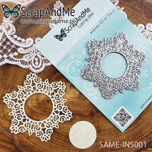 Puede incluir: Un troquel blanco en forma de copo de nieve con una abertura circular en el centro. El troquel está sobre una superficie de madera con un troquel plateado en forma de copo de nieve sobre una tarjeta de fondo azul. La tarjeta tiene el texto "ScrapAndMe" y "www.scrapandme.pl" impreso en ella.