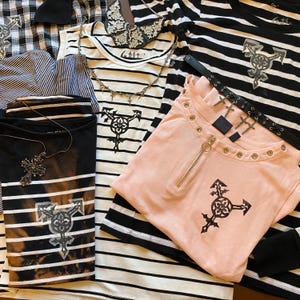 Peut inclure: Un assortiment de vêtements, dont des t-shirts rayés, un haut rose zippé et une chemise à carreaux noirs et blancs. Les chemises présentent des motifs graphiques et sont accessoirisées de colliers et d'une ceinture cloutée.