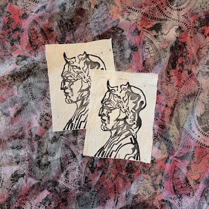 May include: Two small, rectangular art prints featuring a black and white profile of a figure with horns and an ornate headpiece. The prints are on textured paper, set against a colorful paisley fabric background.